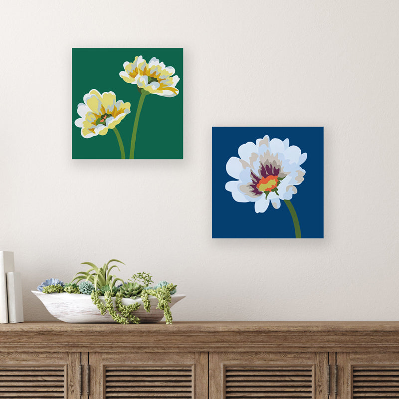 Two 8 inch acrylic and gouache daisy paintings by Michele Aschenbrenner. Two yellow daisies with green background, and a white daisy with blue background displayed on a wall.