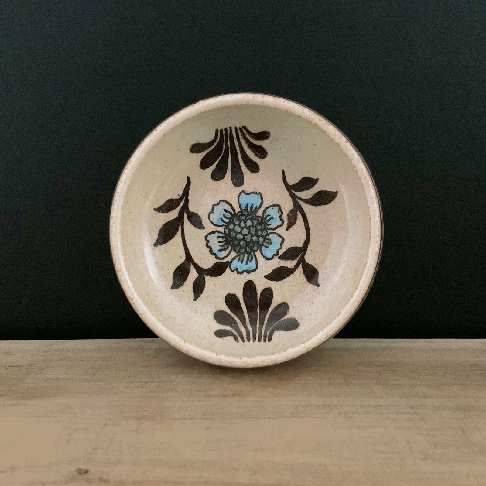 Handmade pottery from Off Menu Pots. Small round catchall dish with blue flower, handmade. Shown with black background.