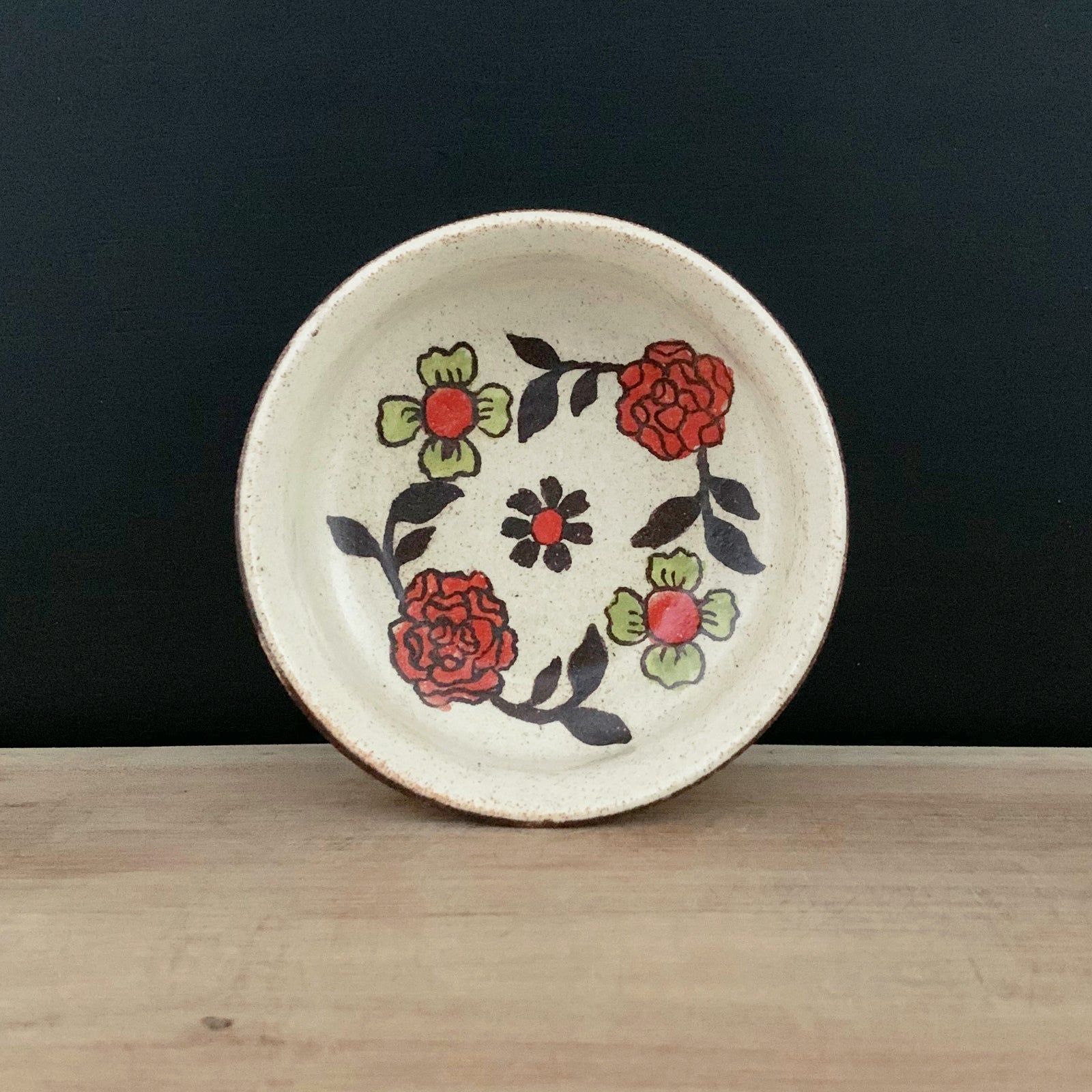 Handmade pottery from Off Menu Pots. Small round catchall dish with red and chartreuse flowers, handmade. Shown with black background.