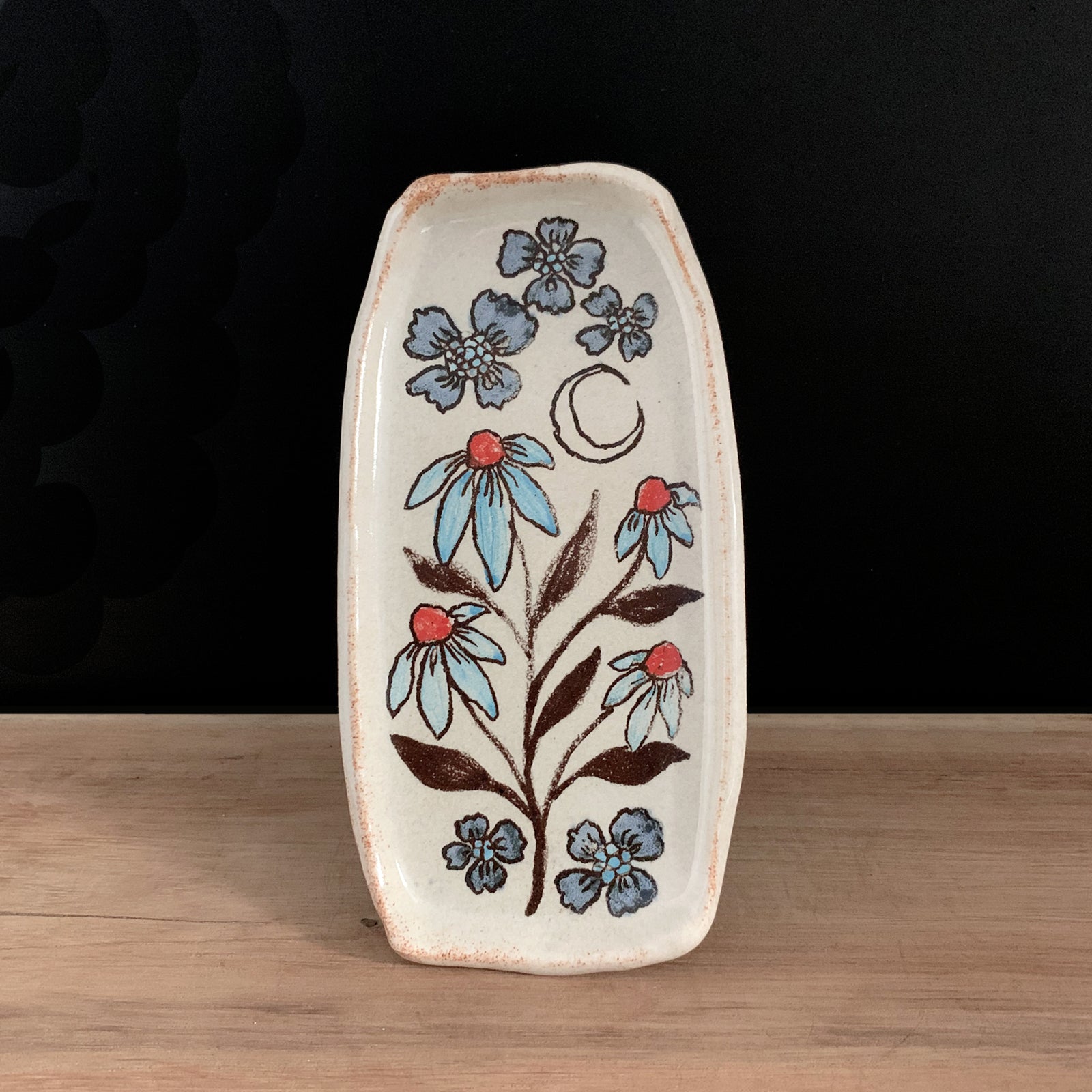 Handmade pottery from Off Menu Pots. Small dish with blue and red flowers, and a crescent moon. Shown on a black background.