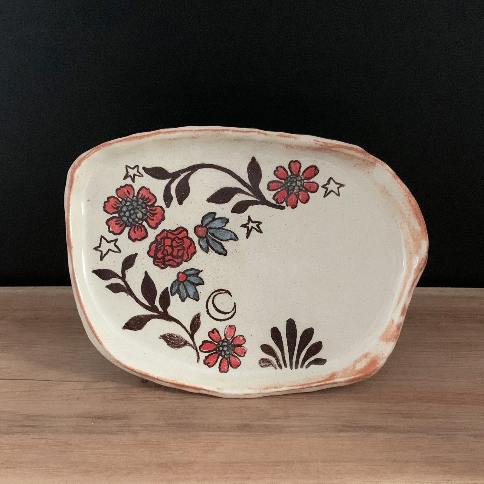 Handmade pottery from Off Menu Pots. Small dish with blue and red flowers, and a crescent moon. Shown with black background.