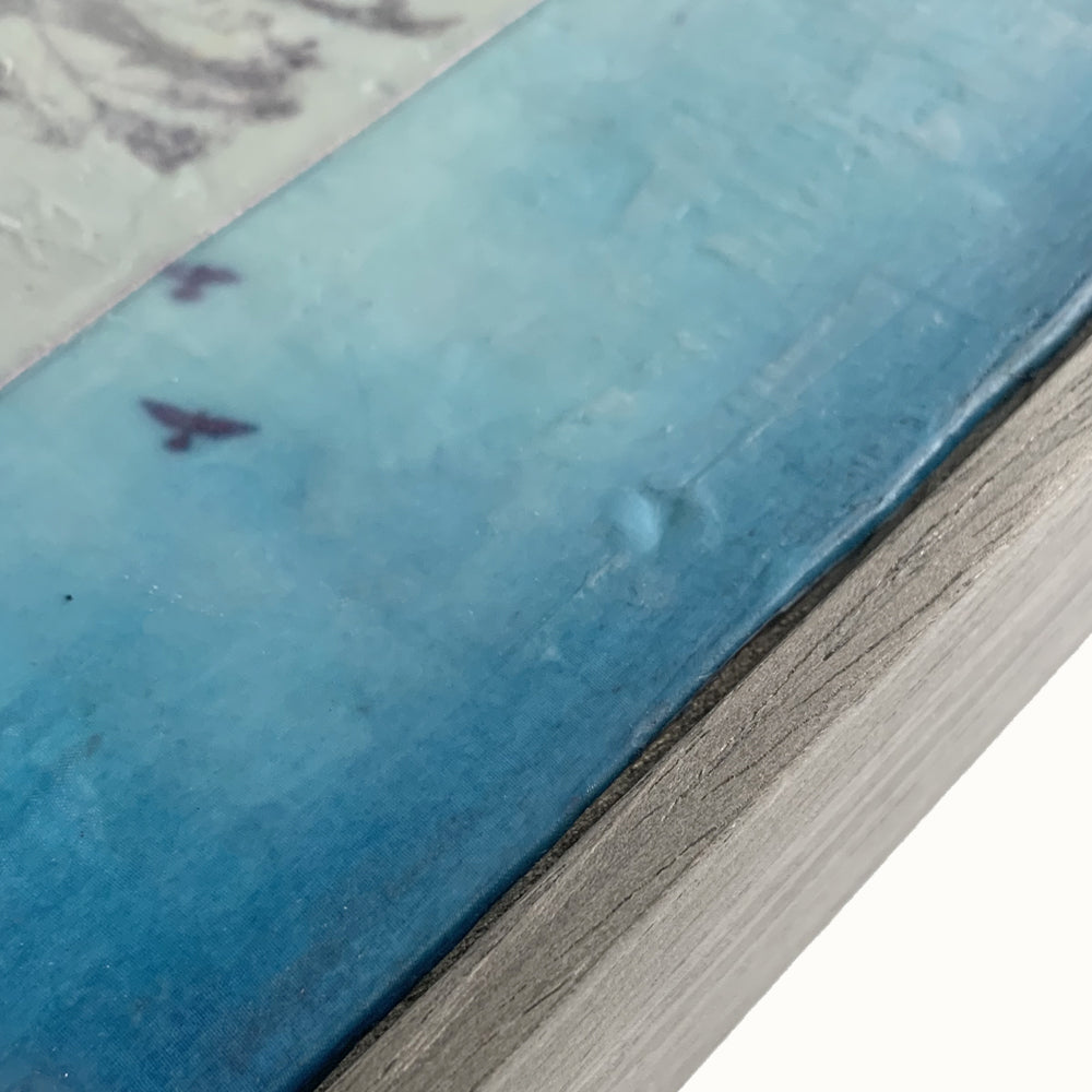 "Surrender" mixed media encaustic painting by Bridgette Guerzon Mills. Framed artwork featuring a textured background, with a tree silhouette on the left and a blue sky with birds on the right. Frame detail shown.