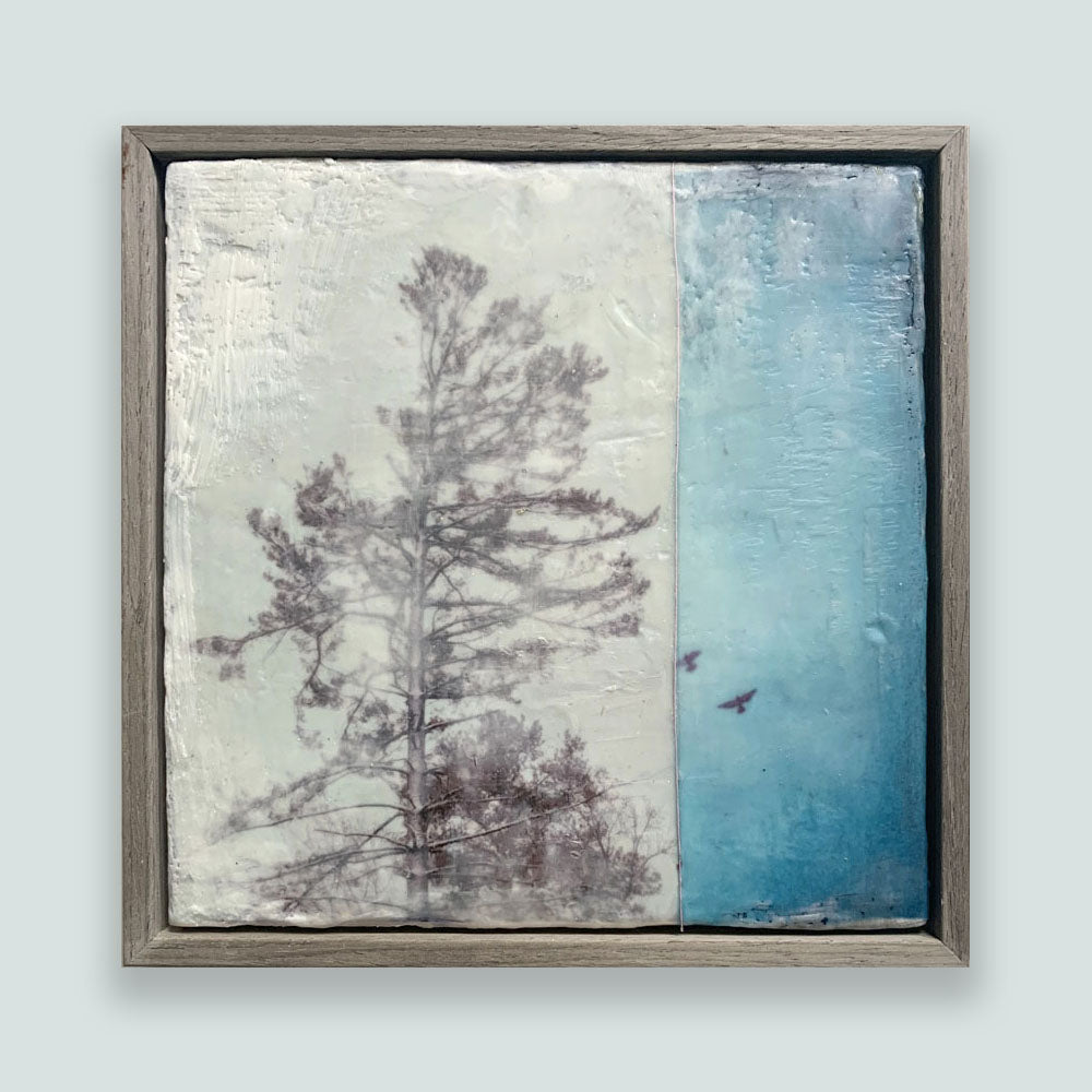 "Surrender" mixed media encaustic painting by Bridgette Guerzon Mills. Framed artwork featuring a textured background, with a tree silhouette on the left and a blue sky with birds on the right. Shown on a pale blue wall.
