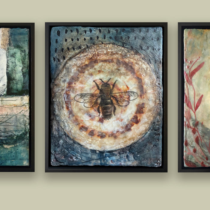 Framed artwork with a bee on a circular design, hung with two other paintings on a beige wall. 
