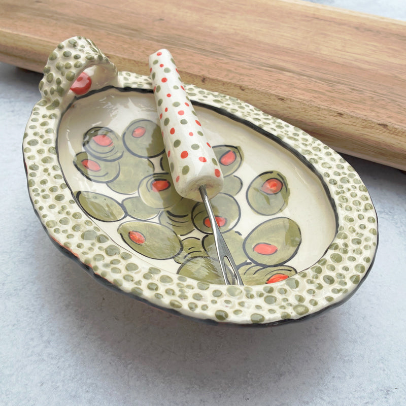 Handmade ceramic olive dish and fork by Molly Lithgo. Textural dish with green olive decoration. Fork with red and green spotted handle. Angled view. 