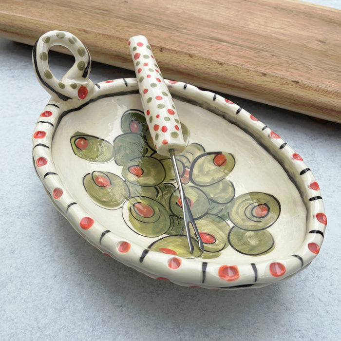 Handmade ceramic olive dish and fork by Molly Lithgo. Oval dish with green olive decoration and polka dots. Fork with red and green spotted handle. Angled view. 