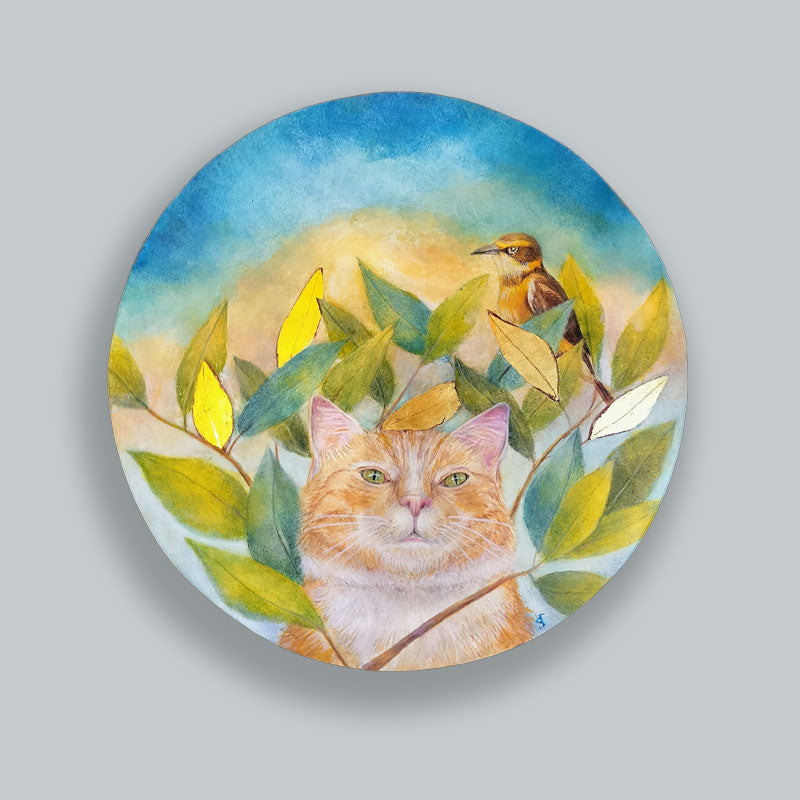A circular egg tempera and gold leaf painting featuring an orange cat looking through a cluster of green leaves, a small bird perched on a branch above, with a soft blue and yellow sky background. Original artwork by Diane Savino displayed on pale blue wall.