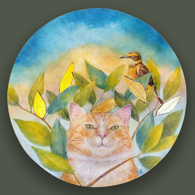 A circular egg tempera and gold leaf painting featuring an orange cat looking through a cluster of green leaves, a small bird perched on a branch above, with a soft blue and yellow sky background. Original artwork by Diane Savino displayed on dark green wall.