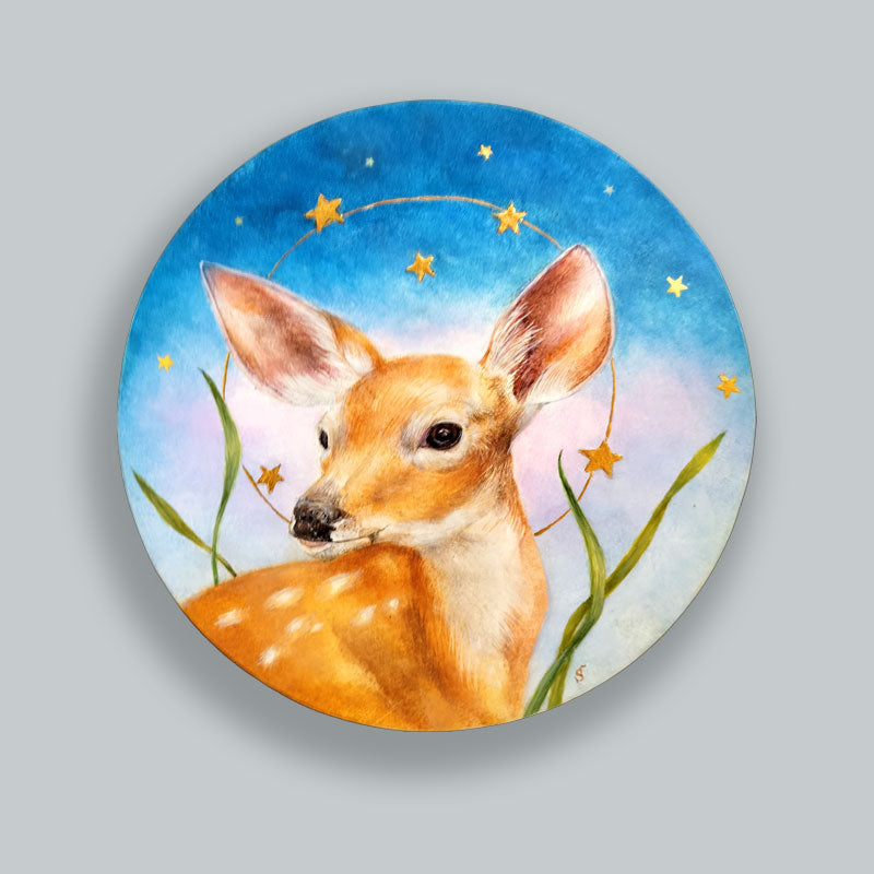 A circular egg tempera and gold leaf painting featuring a young spotted fawn under a starry night sky. Original artwork by Diane Savino, displayed on pale blue wall. 