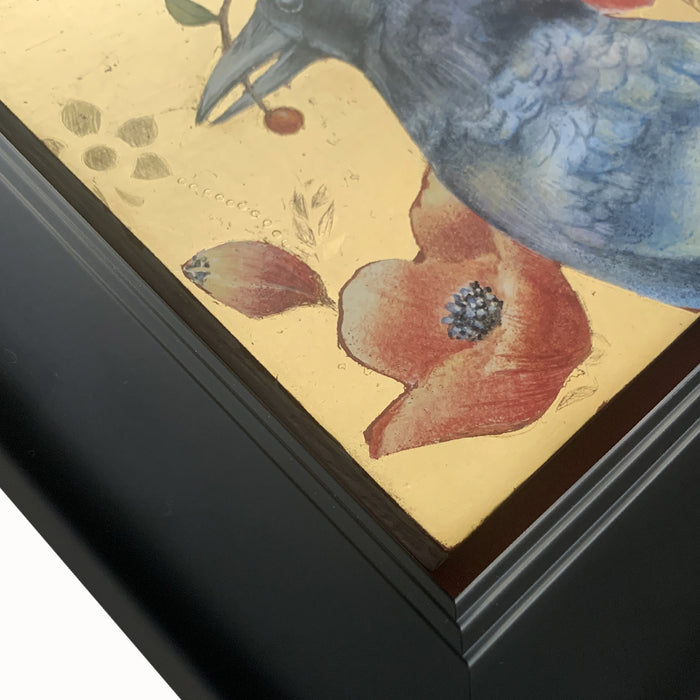"The Bearer" painting by Diane Savino. A crow sits among red flowers with a gold background and black frame. The photo shows a detail of the frame.