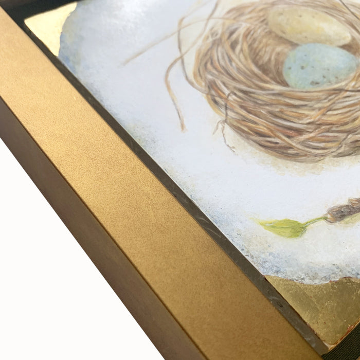 "With Promises to Keep" painting by Diane Savino. A nest holds one blue egg and one yellow egg, a small branch with a single green leaf below it. The artwork has a gold frame, detail shown here.