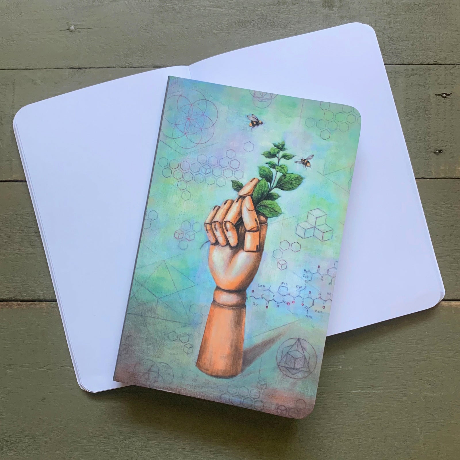 Duy Huynh notebook featuring his "Mint to Bee" artwork, a wooden hand sits among a backdrop of scientific sketches, and holds fresh picked mint with bees flying around it.