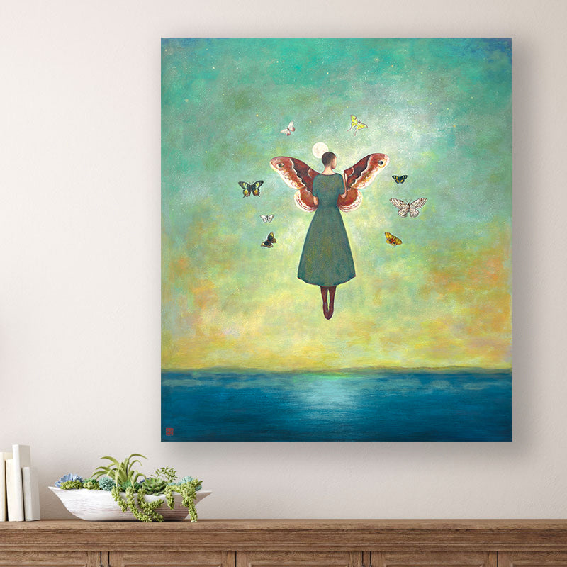 Mythmaker painting by Charlotte NC artist Duy Huynh. Surreal painting of a woman floating above the ocean under a full moon, she is painting large moth wings in the sky and is surrounded by a circle of various moths. The artwork is displayed over a wood console table. 
