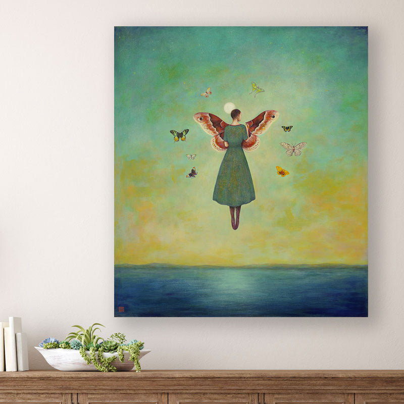 Mythmaker painting by Charlotte NC artist Duy Huynh. Surreal painting of a woman floating above the ocean under a full moon, she is painting large moth wings in the sky and is surrounded by a circle of various moths. The artwork is displayed over a wood console table. 