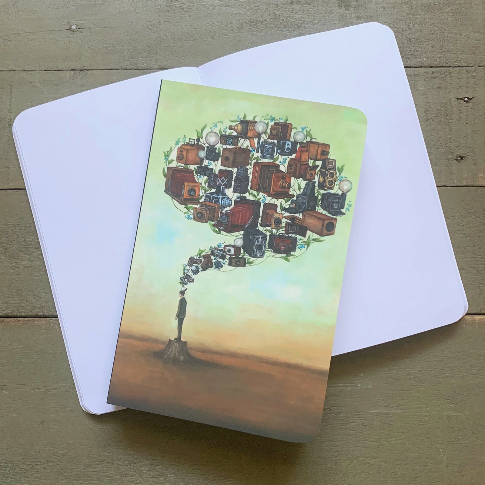 Duy Huynh notebook featuring his "Photographic Memory Brainstem" artwork, a man standing on a tree stump with vintage cameras and forget-me-not flowers rising above into the shape of a brain. Blank notebook pages.