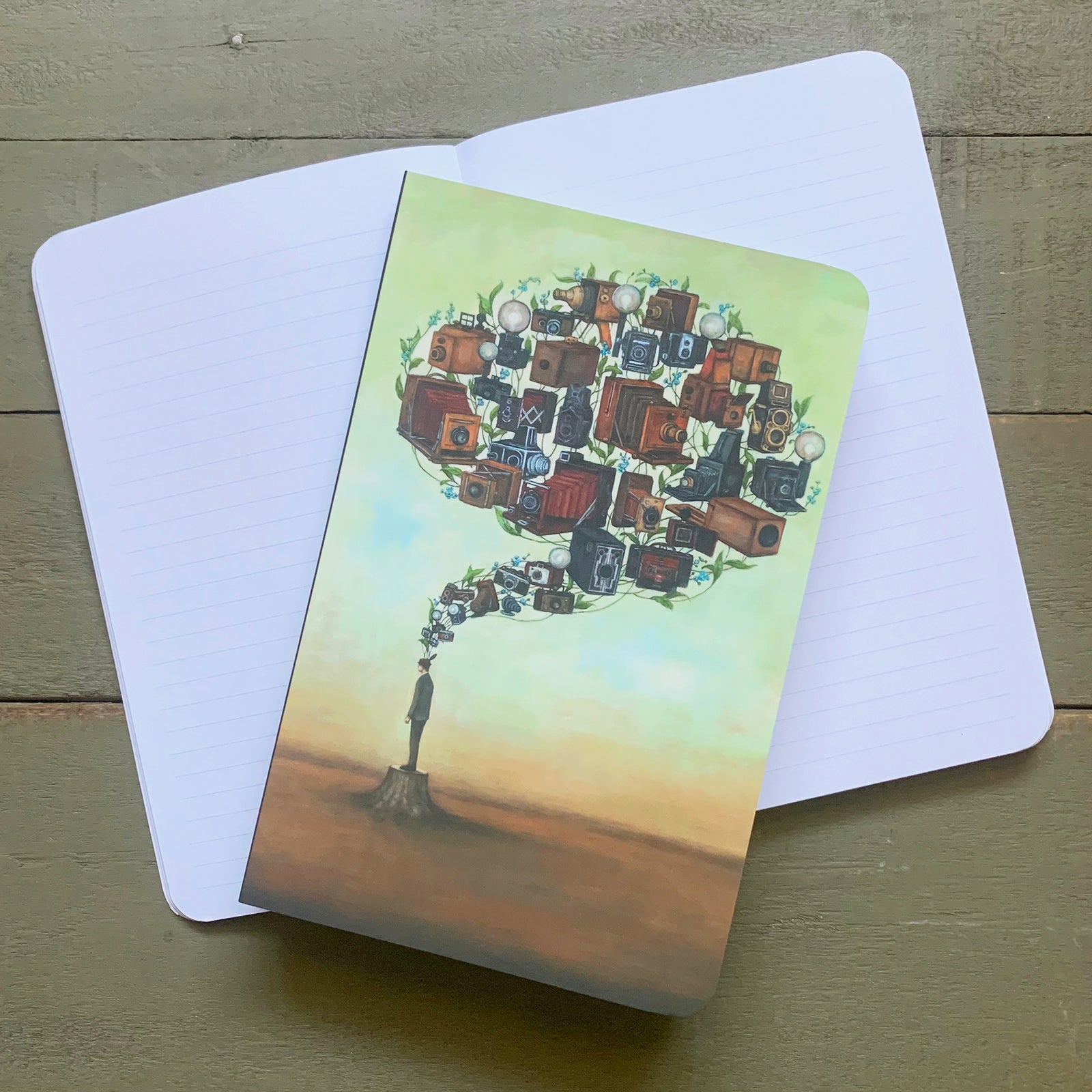Duy Huynh notebook featuring his "Photographic Memory Brainstem" artwork, a man standing on a tree stump with vintage cameras and forget-me-not flowers rising above into the shape of a brain. Lined notebook pages. 