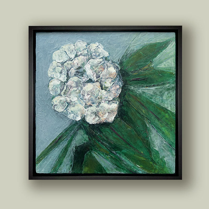 Oil and cold wax painting of a white  flower, green leaves and blue background, framed in black and shown on pale green wall. Artwork by Elizabeth Davant at Lark & Key, Charlotte NC.