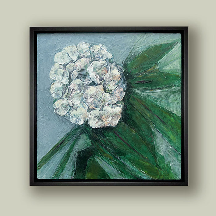 Oil and cold wax painting of a white  flower, green leaves and blue background, framed in black and shown on pale green wall. Artwork by Elizabeth Davant at Lark & Key, Charlotte NC.