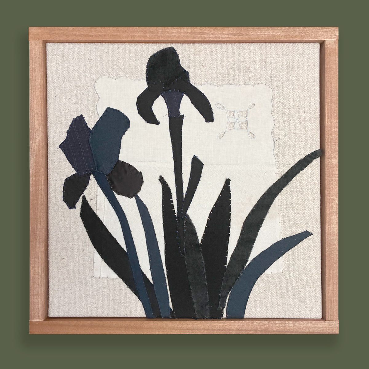 Framed textile artwork of black iris flowers on a textured beige background by Hannah Goff.  Displayed on green wall.