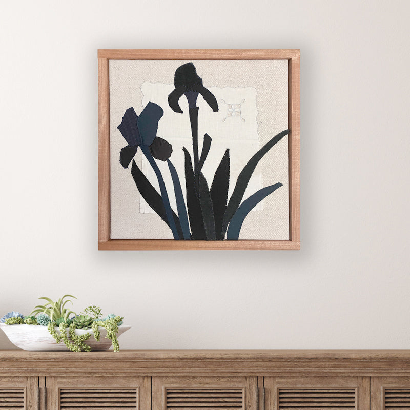 Framed textile artwork of black iris flowers on a textured beige background by Hannah Goff.  Displayed over table. 