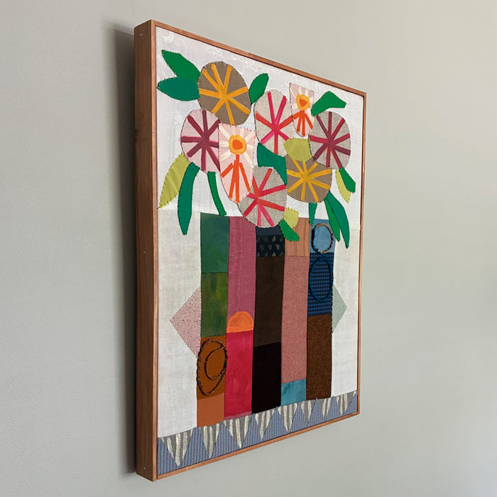 Artwork by Hannah Goff. Mixed media textile painting with geometric vase and colorful abstracted flowers. Side view on wall. 