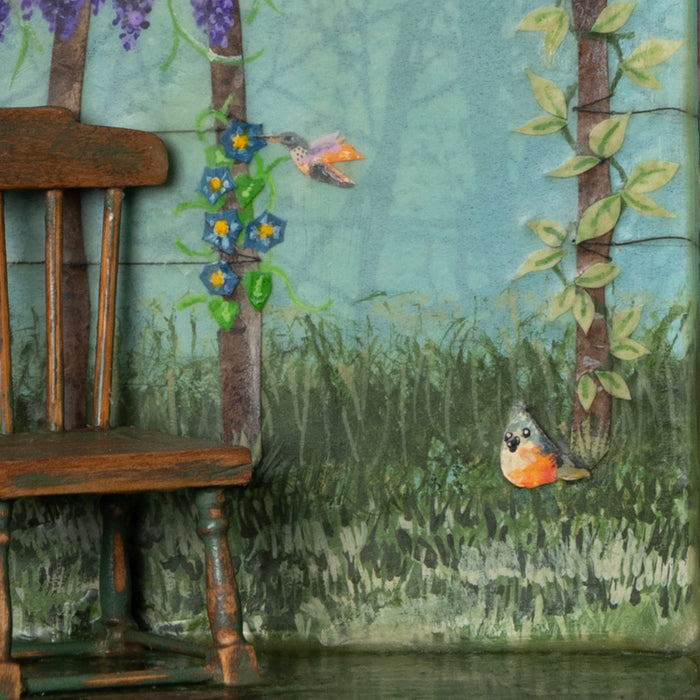 "A Gathering of Feathered Friends", mixed media assemblage inside a vintage wood box with finial on top. The artwork features a blue sky, a variety of birds, flowers on a fence post and a vintage dollhouse chair. By artist Janet Eskridge, close up detail shown.