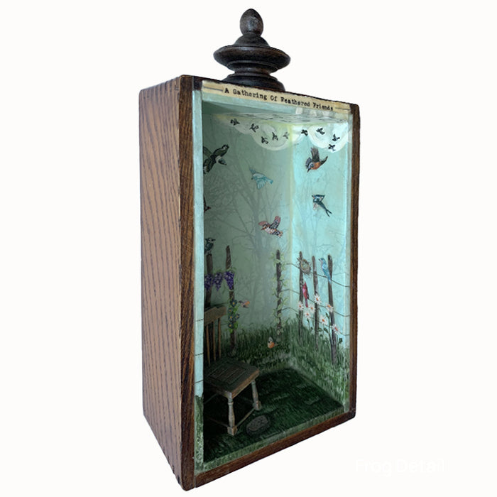 "A Gathering of Feathered Friends", mixed media assemblage inside a vintage wood box with finial on top. The artwork features a blue sky, a variety of birds, flowers on a fence post and a vintage dollhouse chair. Side view showing interior, by artist Janet Eskridge.