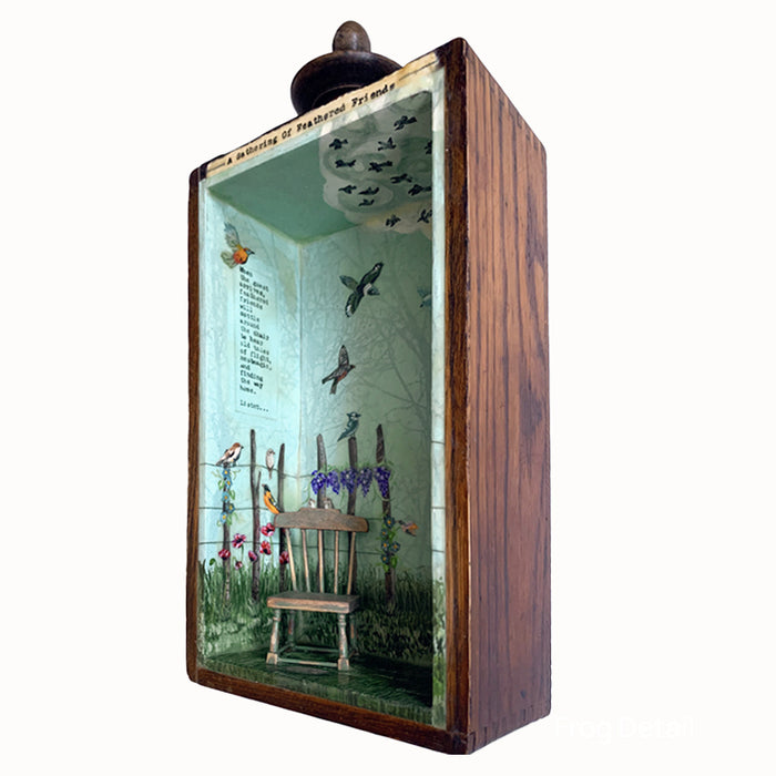 "A Gathering of Feathered Friends", mixed media assemblage inside a vintage wood box with finial on top. The artwork features a blue sky, a variety of birds, flowers on a fence post and a vintage dollhouse chair. Side view showing interior with writing by artist Janet Eskridge.