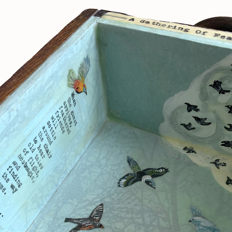 "A Gathering of Feathered Friends", mixed media assemblage inside a vintage wood box with finial on top. The artwork features a blue sky, a variety of birds, flowers on a fence post and a vintage dollhouse chair. Detail with writing by artist Janet Eskridge.