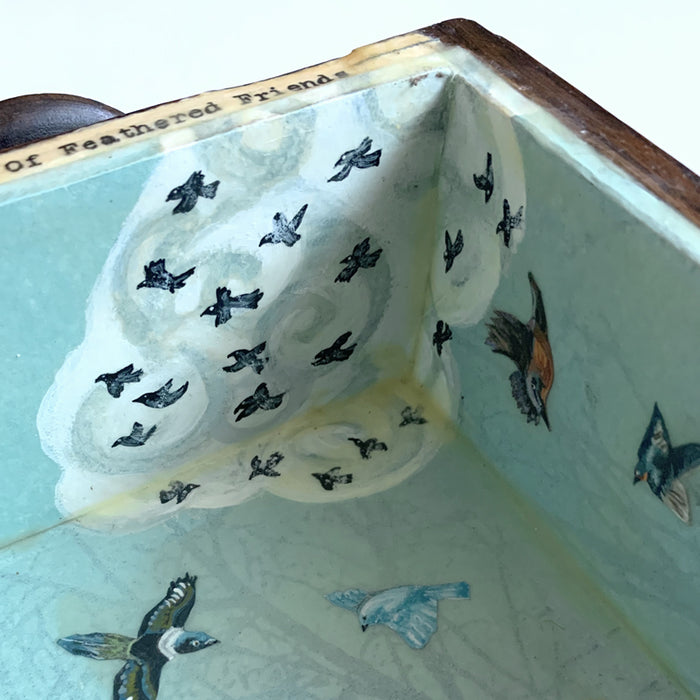 "A Gathering of Feathered Friends", mixed media assemblage inside a vintage wood box with finial on top. The artwork features a blue sky, a variety of birds, flowers on a fence post and a vintage dollhouse chair. Detail of upper interior with birds in the clouds, by artist Janet Eskridge.