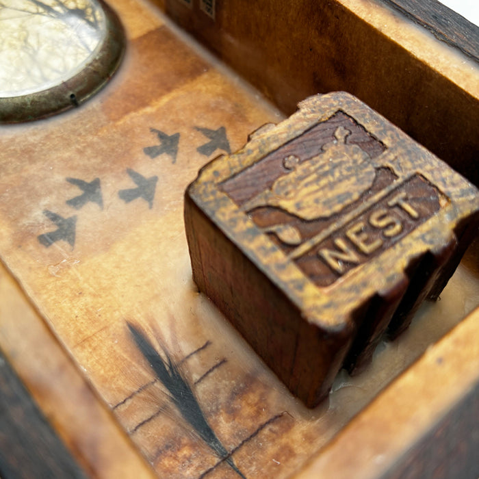 Building a Nest, a mini mixed media assemblage artwork by Janet Eskridge.  Detail of a child's toy block with the word nest, tiny feather, and bird imagery. 