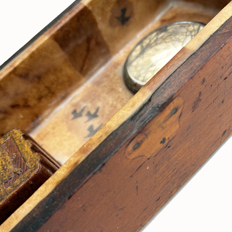 Building a Nest, a mini mixed media assemblage artwork by Janet Eskridge.  A child's toy block with the word nest,  tiny feather,  and tree with nest photo set inside a small round convex clock glass - all set in a vintage wooden box with finial and encased with encaustic. Side detail. 