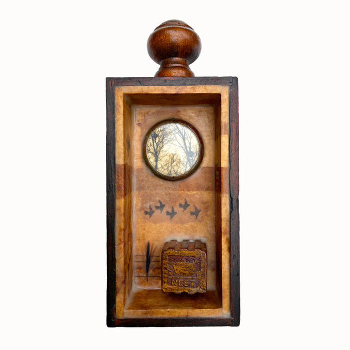 Building a Nest, a mini mixed media assemblage artwork by Janet Eskridge.  A child's toy block with the word nest,  tiny feather,  and tree with nest photo set inside a small round convex clock glass - all set in a vintage wooden box with finial and encased with encaustic. 