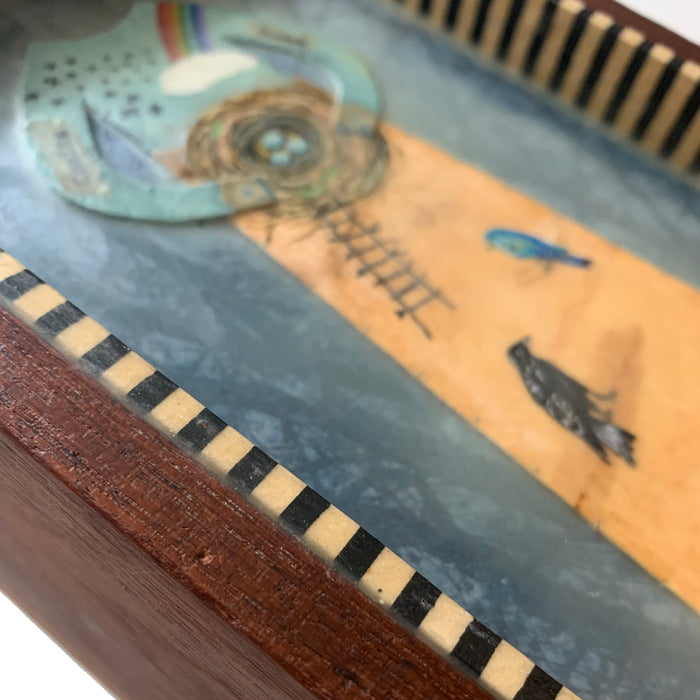 "Homecoming" mixed media assemblage by Janet Eskridge. A figure  in dress holds a bird's next. The dress is decorated with a rainbow, and various birds. The artwork is set inside a vintage wood box with a finial on top. Box detail, side view shown.