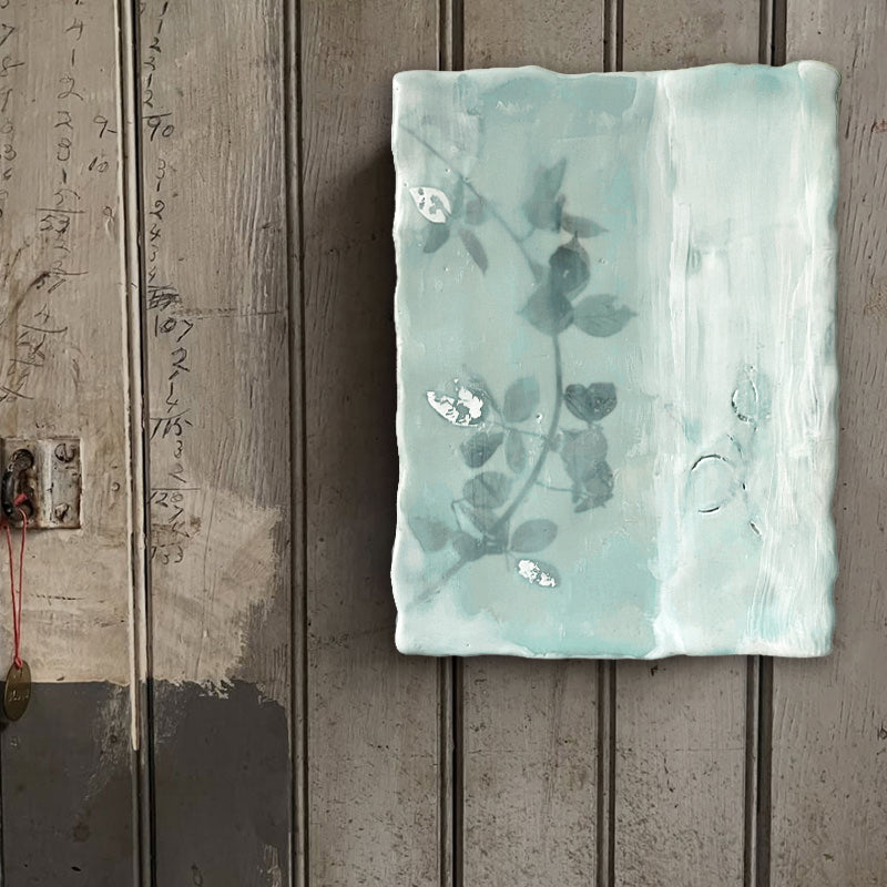 Mixed media encaustic painting by Liberty Hanson Wild in soft green and white with leafy branch and silver leaf detail. Artwork displayed on rustic wood wall. 