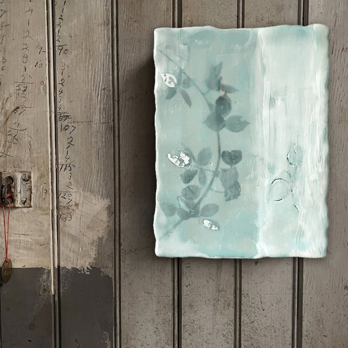 Mixed media encaustic painting by Liberty Hanson Wild in soft green and white with leafy branch and silver leaf detail. Artwork displayed on rustic wood wall. 
