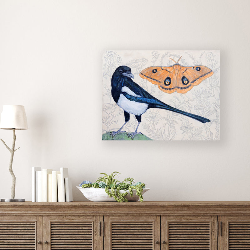 "Magpie Dreams" by artist Martha Worthy. Acrylic painting of a magpie bird and a Polyphemus moth, with a subtle, botanical background. Shown on white wall.