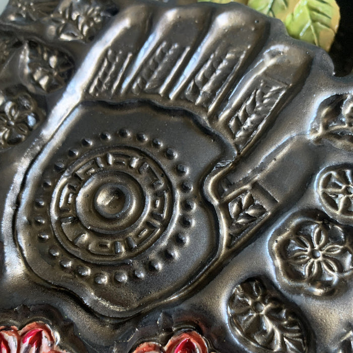 Handmade ceramic wall niche by Paula Smith. Clay face is stamped with flower and bee patterns and a hand pattern, finished in a gunmetal glaze. Red flower details are in the face, with three dimensional yellow light blue flowers with leaves in the center. Hand detail shown.