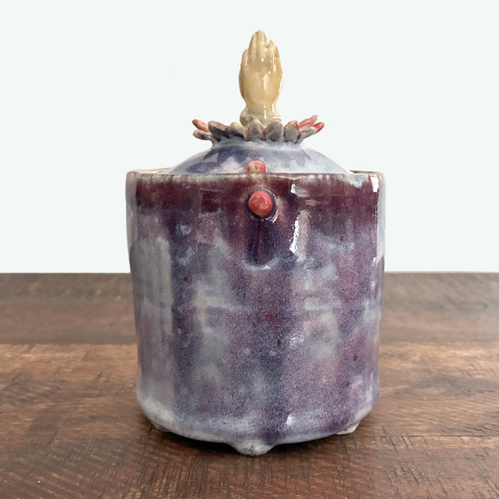 Small purple handmade ceramic jar with lid, lid has flower and hand detail. 