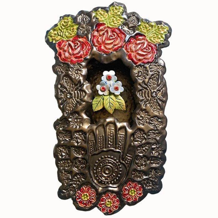 Handmade ceramic wall niche by Paula Smith. Clay face is stamped with flower and bee patterns and a hand pattern, finished in a gunmetal glaze. Red flower details are in the face, with three dimensional yellow light blue flowers with leaves in the center. 