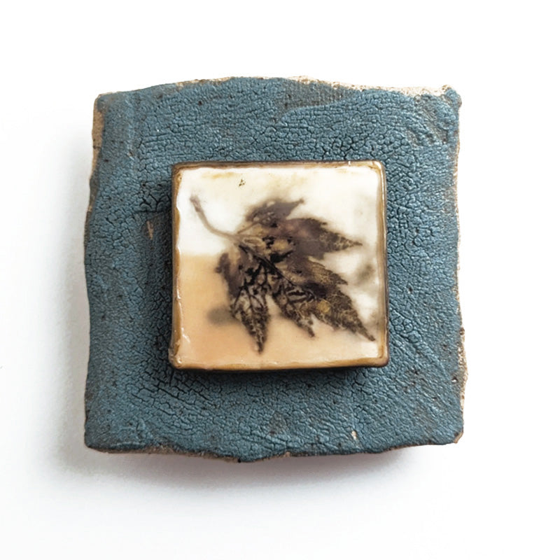 Small ceramic tile with textured blue glaze, with a botanical print encased in wax set in the center. Artwork by Sarah Treanor. 