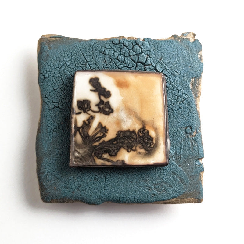 Small ceramic tile with textured blue glaze, with a botanical print encased in wax set in the center. Artwork by Sarah Treanor. 