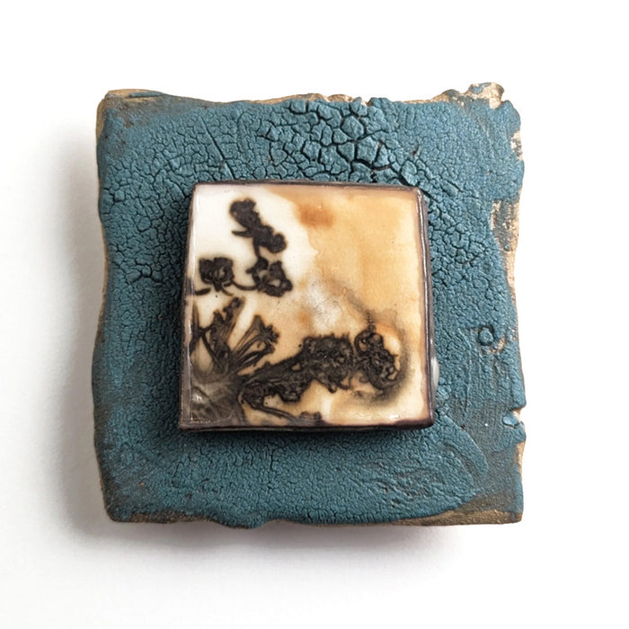 Small ceramic tile with textured blue glaze, with a botanical print encased in wax set in the center. Artwork by Sarah Treanor. 