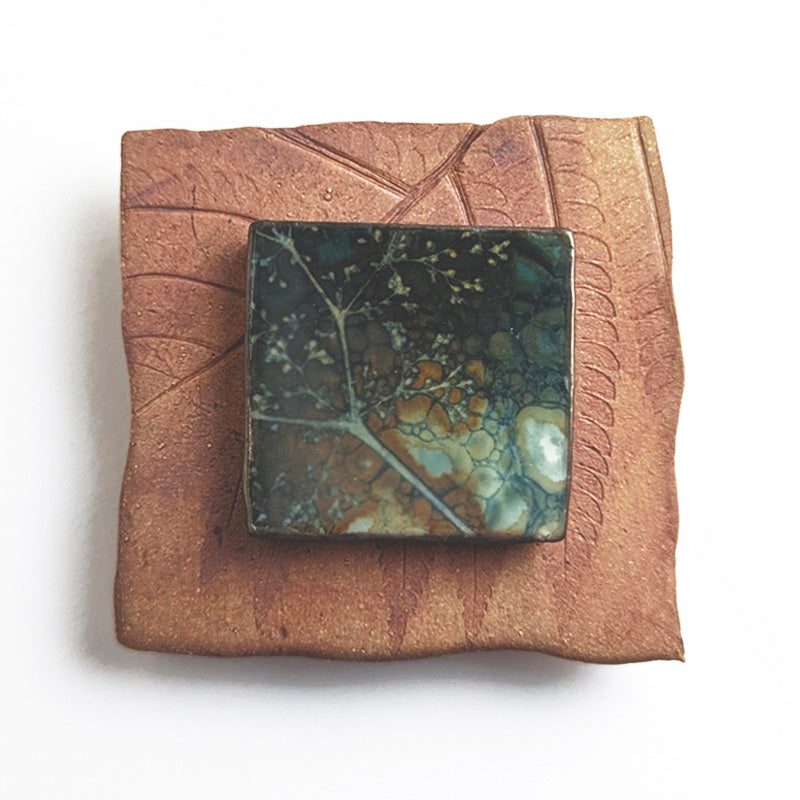 Small ceramic tile with textured blue glaze, with a botanical print encased in wax set in the center. Artwork by Sarah Treanor. 