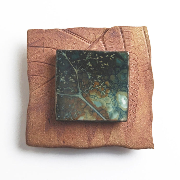 Small ceramic tile with textured blue glaze, with a botanical print encased in wax set in the center. Artwork by Sarah Treanor. 