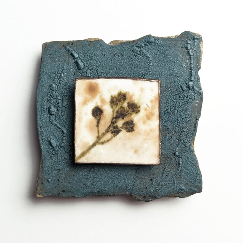 Small ceramic tile with textured blue glaze, with a botanical print encased in wax set in the center. Artwork by Sarah Treanor. 