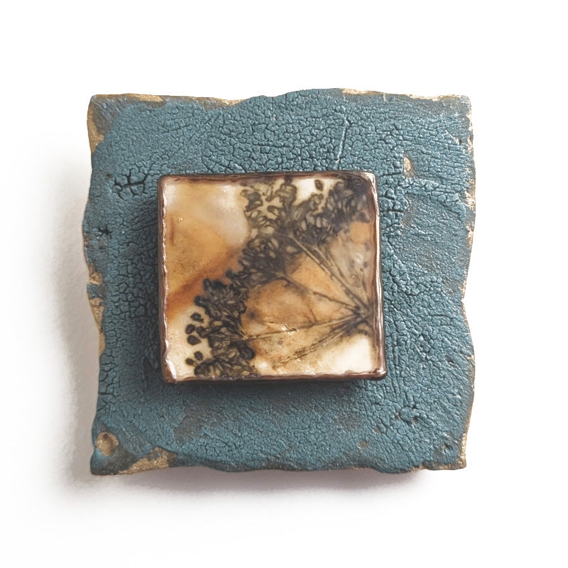 Small ceramic tile with textured blue glaze, with a botanical print encased in wax set in the center. Artwork by Sarah Treanor. 