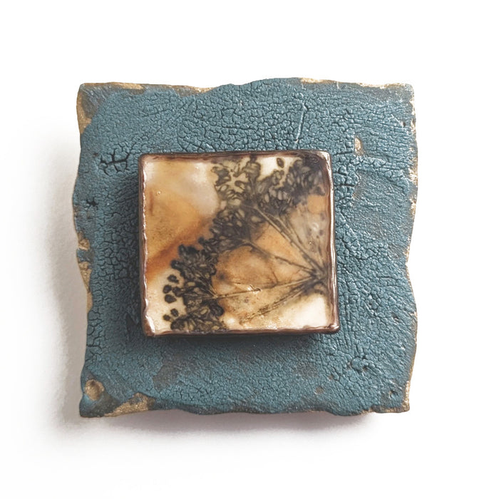Small ceramic tile with textured blue glaze, with a botanical print encased in wax set in the center. Artwork by Sarah Treanor. 