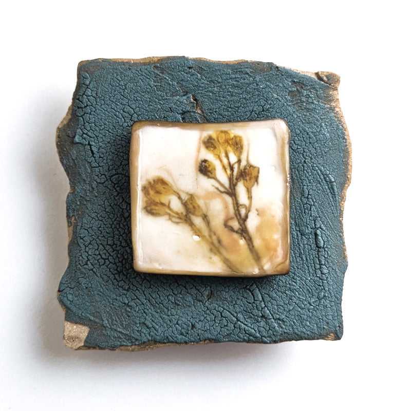 Small ceramic tile with textured blue glaze, with a botanical print encased in wax set in the center. Artwork by Sarah Treanor. 