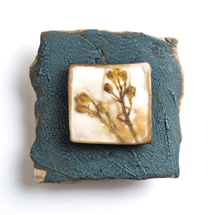 Small ceramic tile with textured blue glaze, with a botanical print encased in wax set in the center. Artwork by Sarah Treanor. 
